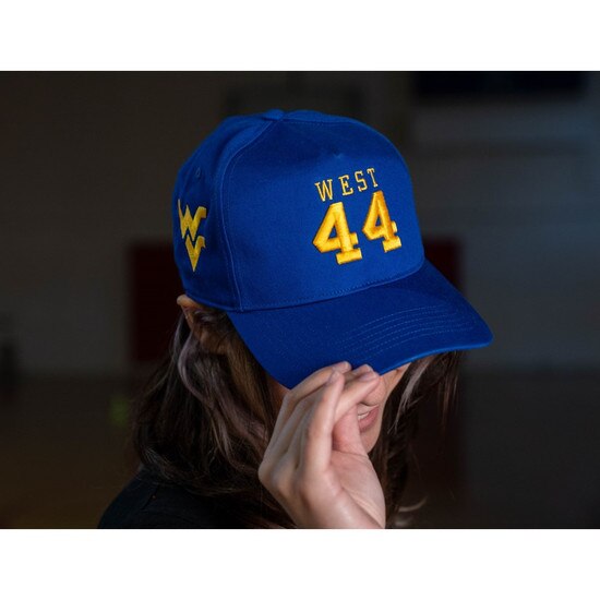 Model wearing the royal blue WVU Jerry West hat with West over 44 embroidered in gold on the front panels and a gold Flying WV embroidered on the lower left panels