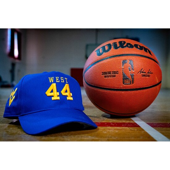 The royal blue WVU Jerry West hat with West over 44 embroidered in gold on the front panels and a gold Flying WV embroidered on the lower left panel sitting on a basketball court next to a basketball