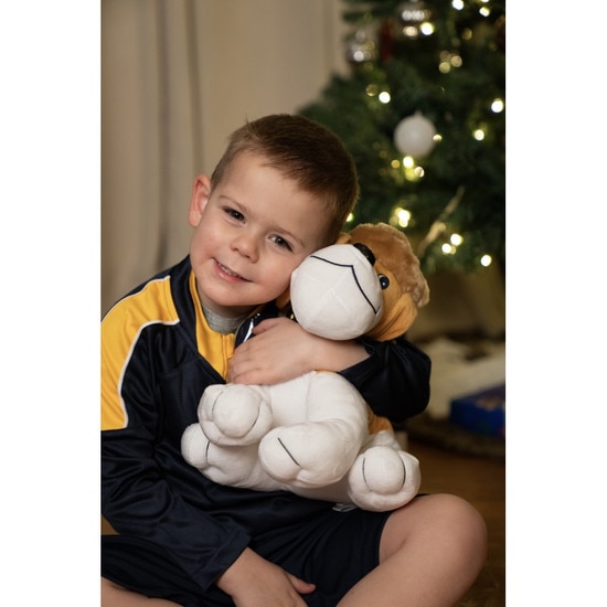 A child holding the cute WVU 11" tall Musket the Dog plushie wearing a Mountaineer coon skin hat and a WVU collar while sitting in front of a Christmas tree